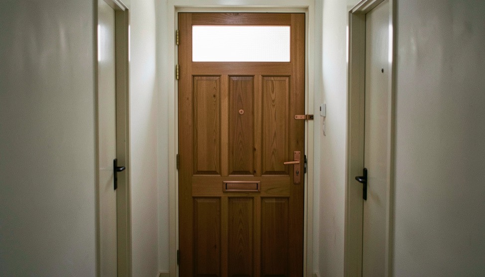 Een gesloten houten deur met een koperen klink en een matglazen bovenlicht in een Nederlandse woninggang.